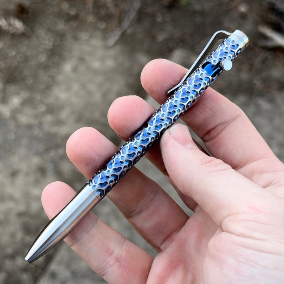 A hand holding a metallic pen with a textured, blue and silver design on the barrel. The background is blurred outdoor ground.