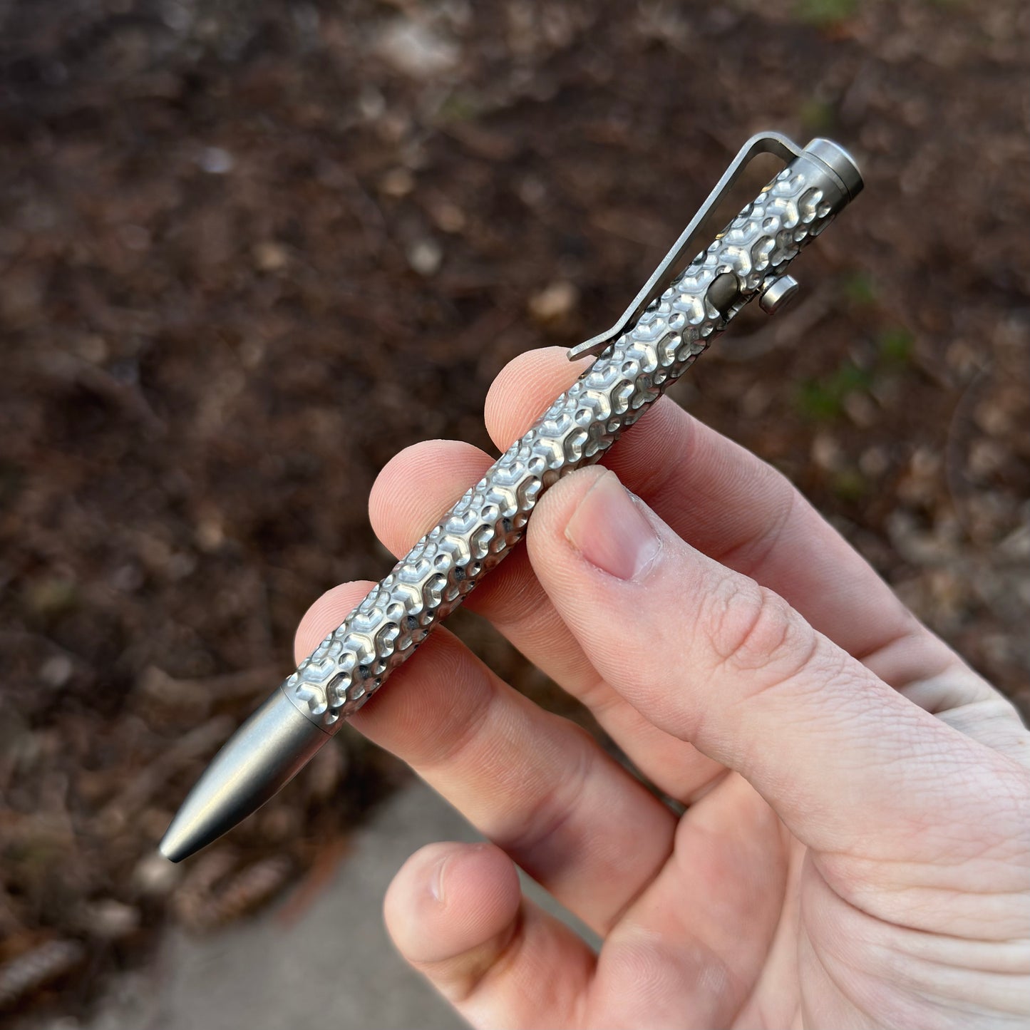 Close-up of a hand holding a textured metal pen with a clip against a blurred outdoor background.