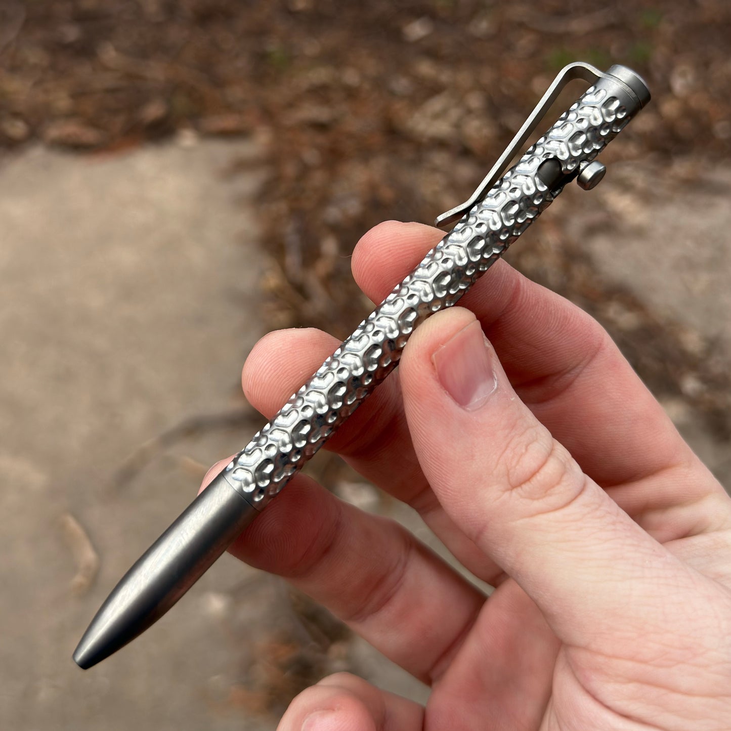 Person holding a textured metal pen with a tactile grip and sleek design on an outdoor cement pathway background.