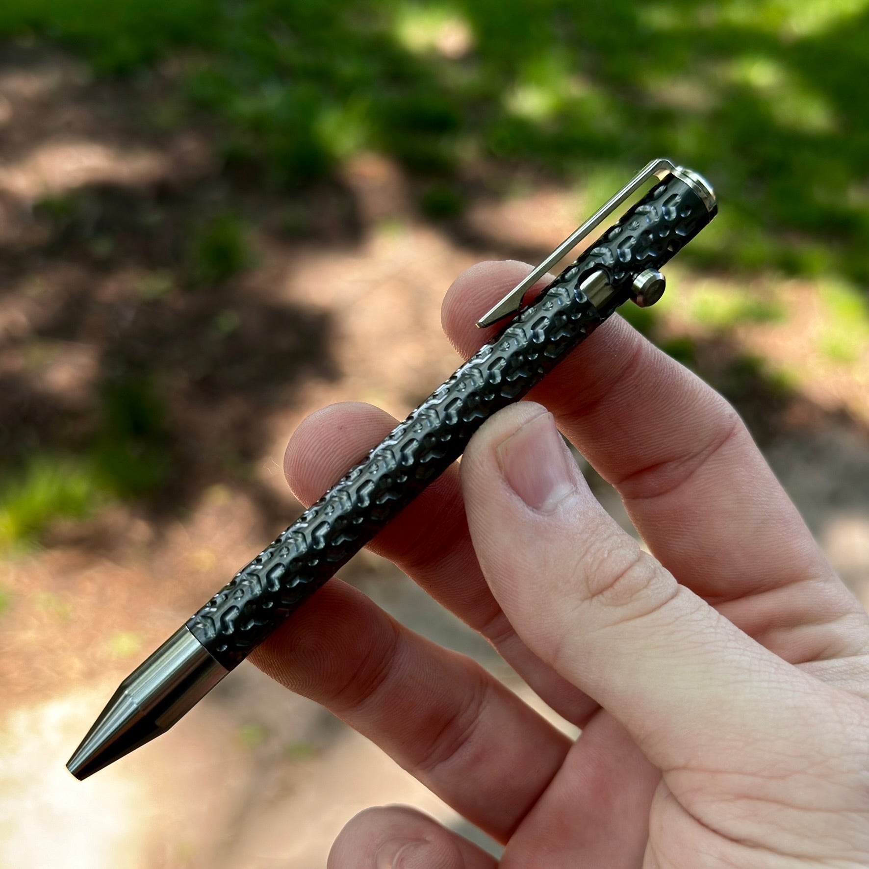 Hand holding a black and silver patterned tactical pen with a clip, outdoors with blurred green and brown background.